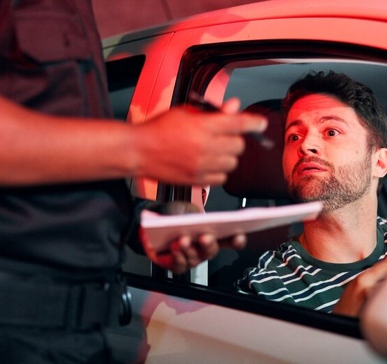 Driver looks worried as a police officer writes a citation during a nighttime traffic stop, highlighting most common traffic violations that raise insurance rates.