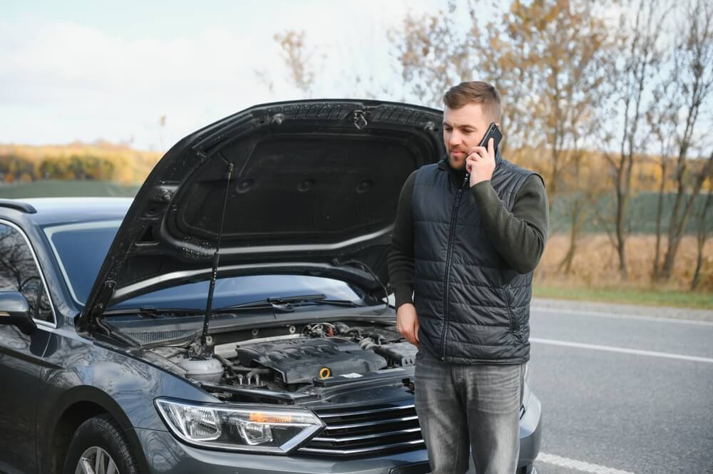 Man calls for help beside a car with the hood open on the side of the road, showing how roadside assistance coverage can support drivers during a breakdown.
