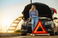 Woman waits beside a broken-down car with the hood open and a warning triangle nearby, showing why roadside assistance coverage can be helpful during unexpected stops.