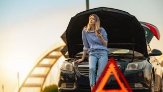 Woman waits beside a broken-down car with the hood open and a warning triangle nearby, showing why roadside assistance coverage can be helpful during unexpected stops.