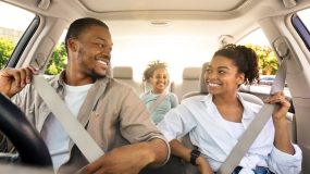 Smiling family of three rides in a car with everyone buckled up, illustrating seat belt safety laws.