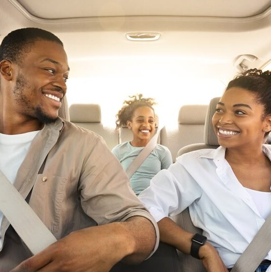 Smiling family of three rides in a car with everyone buckled up, illustrating seat belt safety laws.