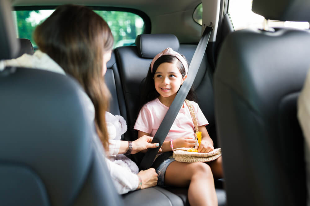 Adult woman helps a young girl buckle her seat belt in the back seat of a car, reinforcing seat belt safety laws.
