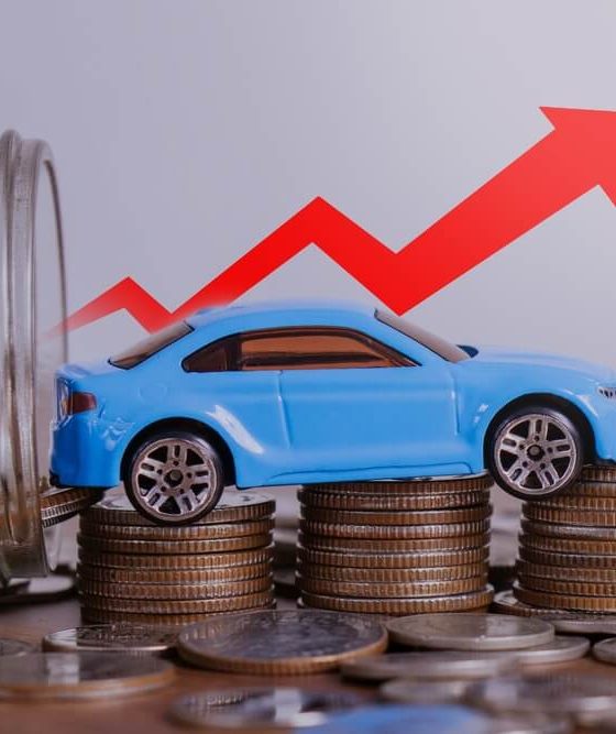 Blue toy car sits on stacks of coins beside a jar of change, with a red arrow pointing upward to show costs rising and car insurance going up.