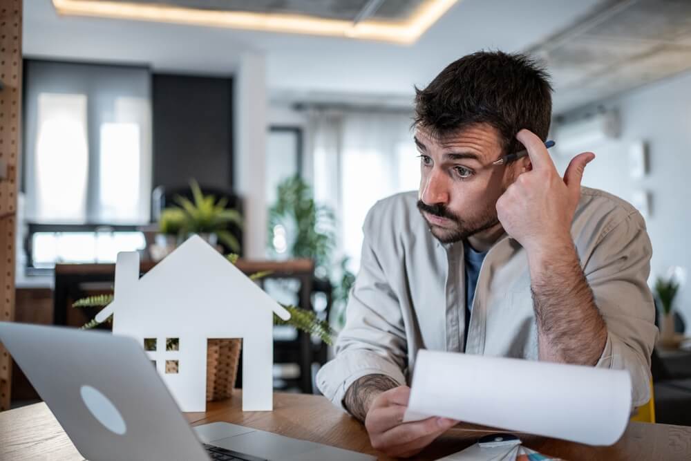 Man reviewing bills beside a house model, reflecting financial concerns and need for homeowners insurance cover protection