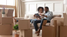 Young couple sitting on the couch amongst a stack of moving boxes looking at digital tablet and taking a break