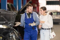 Mechanic in blue overalls talks with a concerned woman beside a car with the hood open in a repair shop, illustrating best times to take your car to the auto shop.