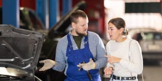 Mechanic in blue overalls talks with a concerned woman beside a car with the hood open in a repair shop, illustrating best times to take your car to the auto shop.