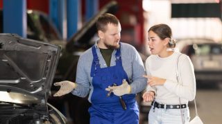 Mechanic in blue overalls talks with a concerned woman beside a car with the hood open in a repair shop, illustrating best times to take your car to the auto shop.