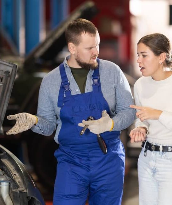 Mechanic in blue overalls talks with a concerned woman beside a car with the hood open in a repair shop, illustrating best times to take your car to the auto shop.