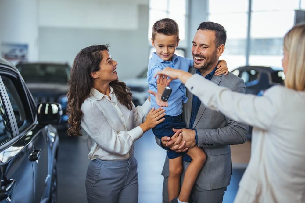 Happy family in dealership showroom accepts keys to new car - cheap car insurance.