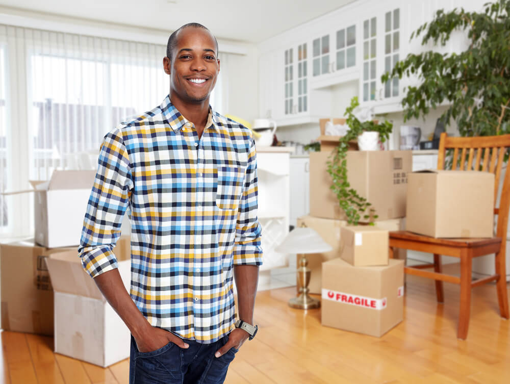 Young black man stands in the living room of his condo with unpacked boxes around - cheap condo insurance.