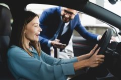 Very happy young woman sitting behind the wheel of her new car with the car salesman standing outside, deciding whether to lease or buy her new vehicle.