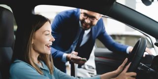 Very happy young woman sitting behind the wheel of her new car with the car salesman standing outside, deciding whether to lease or buy her new vehicle.