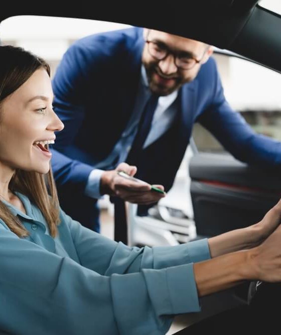 Very happy young woman sitting behind the wheel of her new car with the car salesman standing outside, deciding whether to lease or buy her new vehicle.