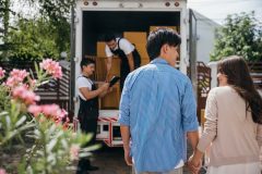 Assisted by a professional delivery team a couple transitions to their new home. Together they unload and lift cardboard boxes ensuring an organized and efficient move to their new rental home with transferred renters insurance.
