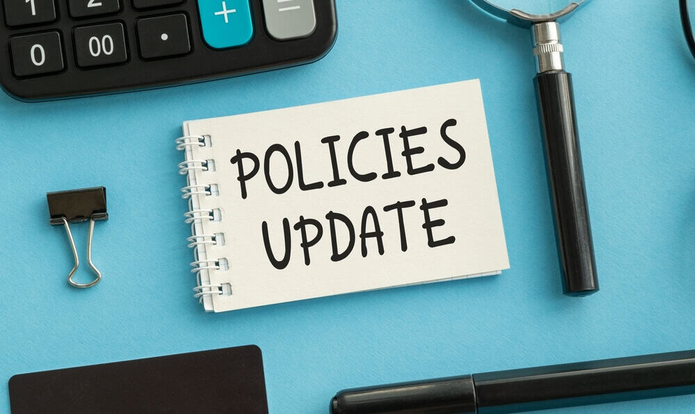 Blue background, spiral notebook with words "Policies Update", binder clip, marker and magnifying glass to describe concept of renewing your insurance policy.