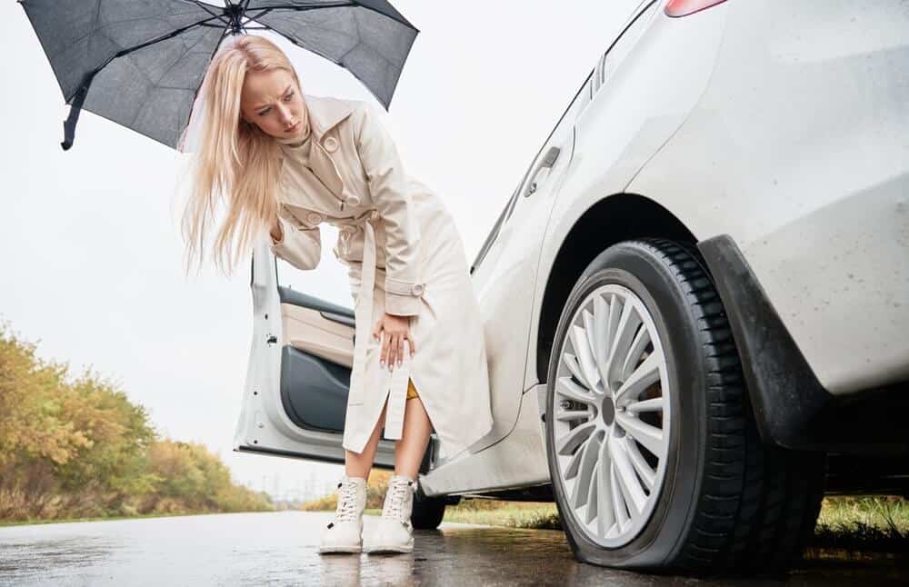 Woman on a road near her white car with punctured car tire, holding umbrella in the rain, need for cheap roadside assistance to come and change tire.