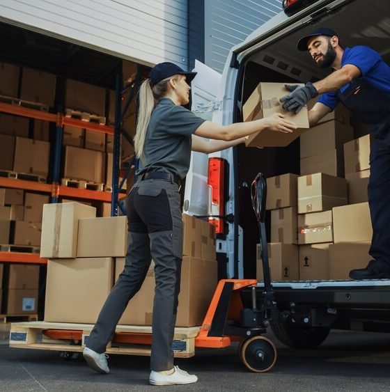 Outside of logistics distributions warehouse: diverse team of workers use hand truck loading delivery van with cardboard boxes, online orders, e-commerce purchases. Concept of when commercial auto insurance is right for you.