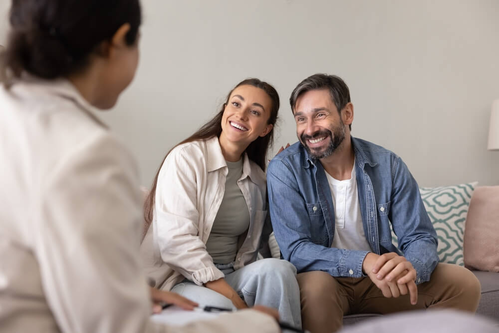 A smiling couple meets with an insurance agent at home, learning how to avoid common auto insurance mistakes.