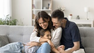 Happy young parents cuddling their daughter on the living-room couch, illustrating how avoiding Covered California enrollment mistakes helps protect their family’s health coverage