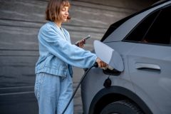 Young woman charging her electric vehicle at home while using her phone, illustrating everyday situations covered by electric vehicle insurance.