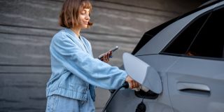 Young woman charging her electric vehicle at home while using her phone, illustrating everyday situations covered by electric vehicle insurance.