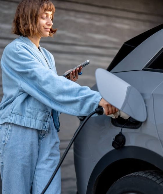 Young woman charging her electric vehicle at home while using her phone, illustrating everyday situations covered by electric vehicle insurance.
