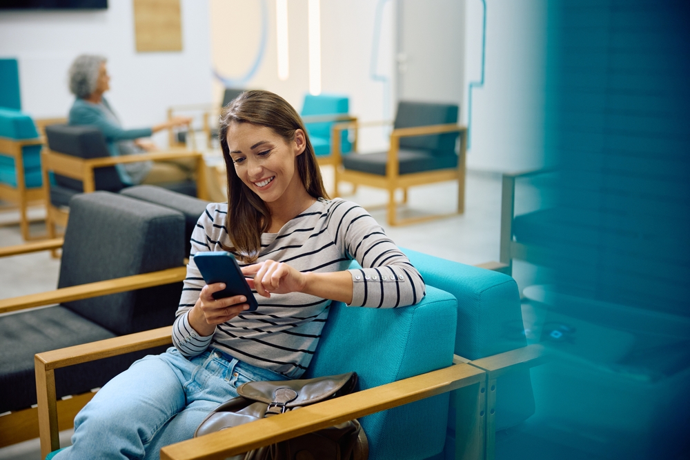 Smiling woman sits in waiting room at doctor's office, using her phone to check her Covered California 2026 benefits.