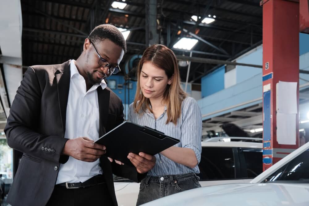 Man in a suit and glasses reviews a clipboard with a woman in a striped shirt at an auto repair shop, as they go over reasons for car insurance going up.