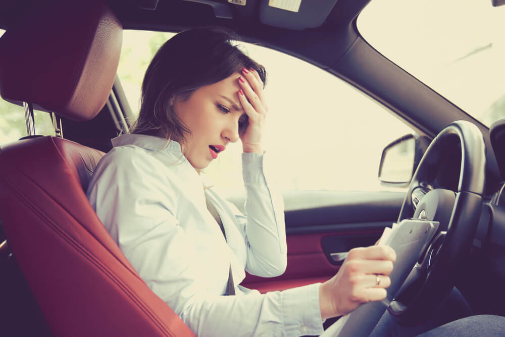 Stressed woman in a white button-up shirt sits in the driver’s seat with her hand on her forehead while reading a bill or paperwork, reacting to car insurance going up.