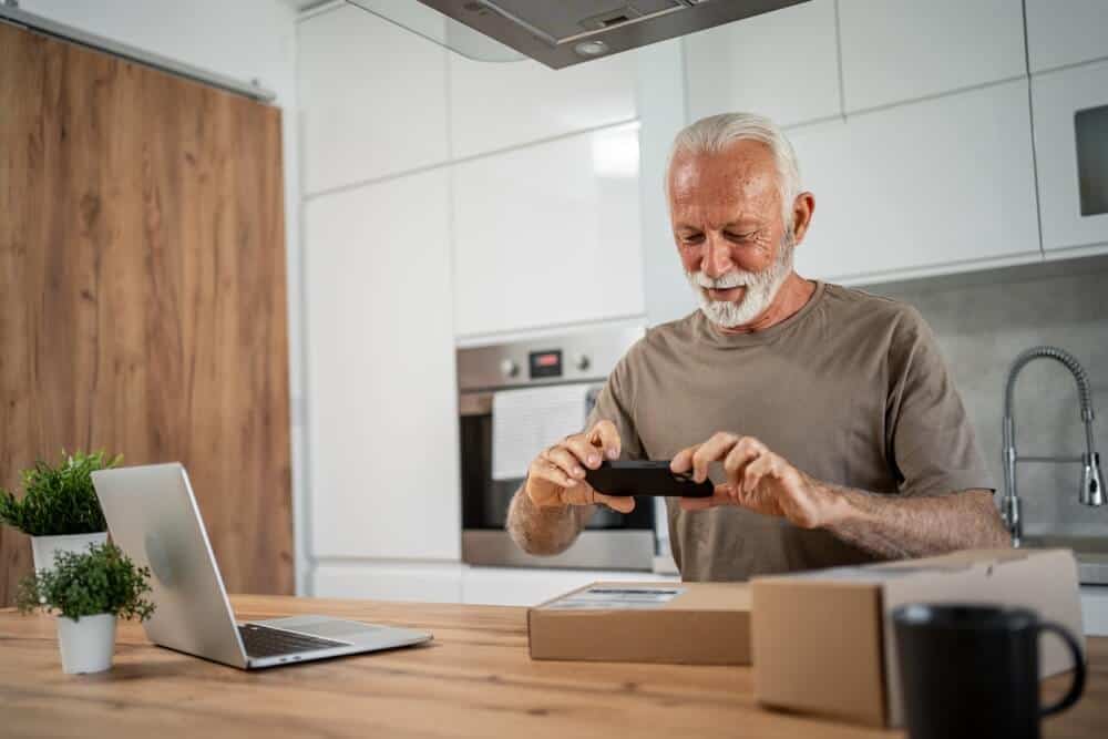 Happy retired man taking pictures of his package with smartphone in modern kitchen, sitting by the table with laptop and cup of coffee or tea near him.