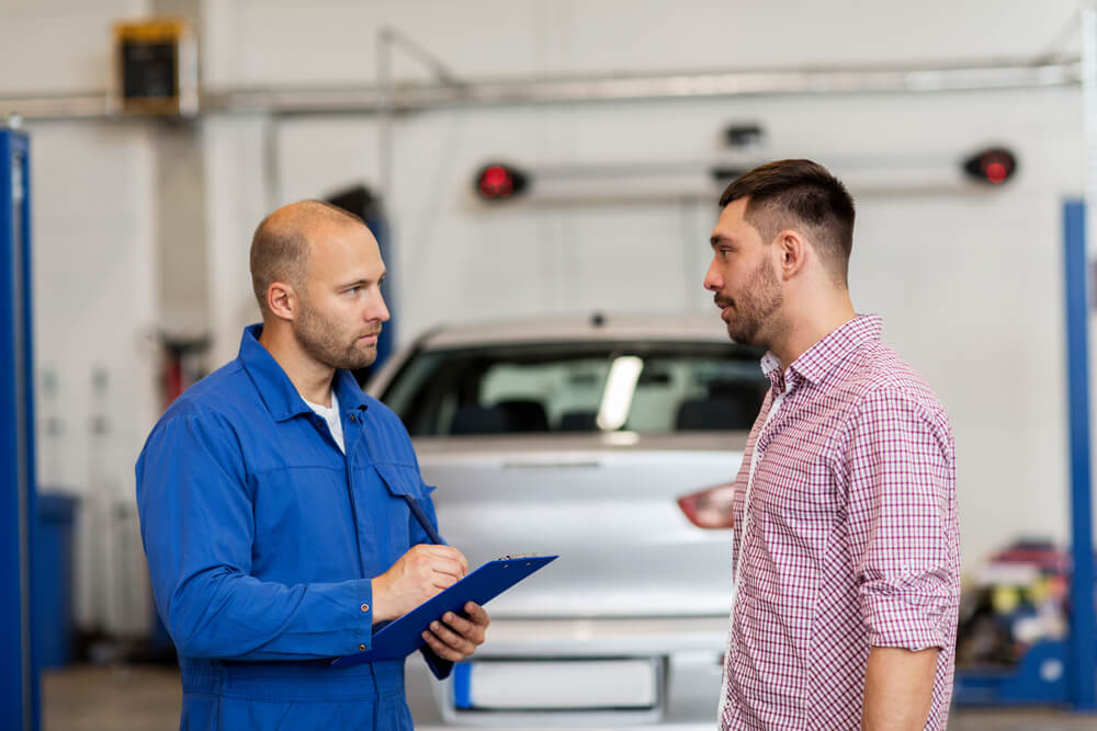 Auto mechanic in blue coveralls writes on a clipboard while talking with a customer in a repair bay, a reminder of the best times to take your car to the auto shop.