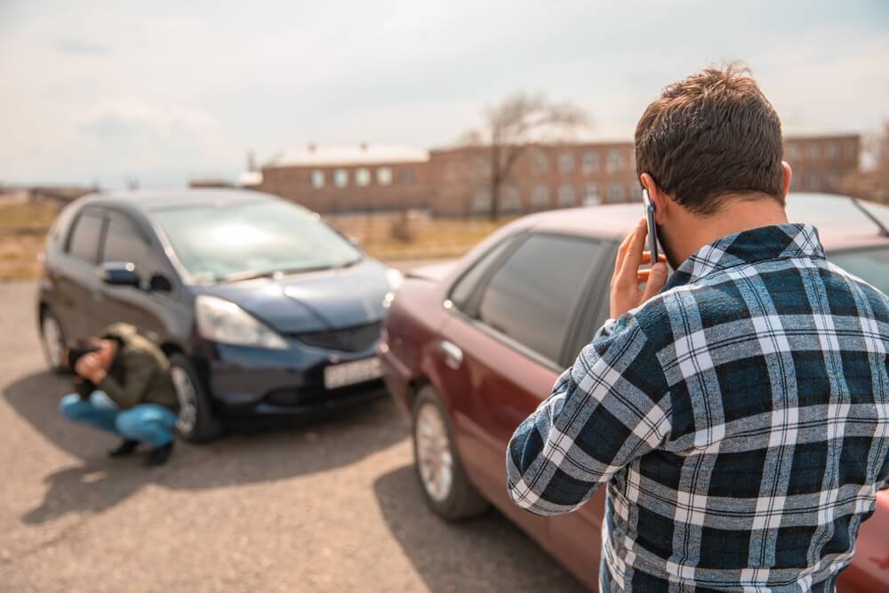 Driver calling his provider for insurance after an accident, standing beside a damaged car while another person inspects the front bumper after a minor collision in a parking lot.
