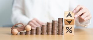 Rising stacks of coins beside a toy car and blocks with arrows and a percent symbol representing why car insurance rates change, including factors like inflation, accident claims, repair costs, driver risk, and insurance market trends.