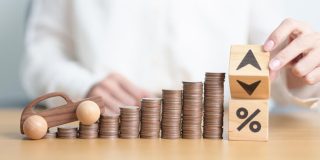 Rising stacks of coins beside a toy car and blocks with arrows and a percent symbol representing why car insurance rates change, including factors like inflation, accident claims, repair costs, driver risk, and insurance market trends.