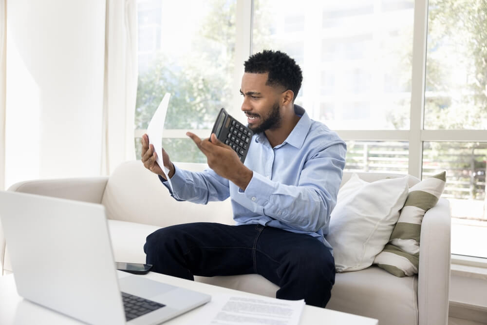 Man sitting on a couch looks stressed while holding a calculator and paperwork beside a laptop, illustrating what happens if insurance lapses.