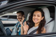 Smiling driver and passenger make “OK” hand signs inside a car, highlighting permissive driving.