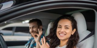 Smiling driver and passenger make “OK” hand signs inside a car, highlighting permissive driving.