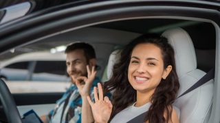 Smiling driver and passenger make “OK” hand signs inside a car, highlighting permissive driving.