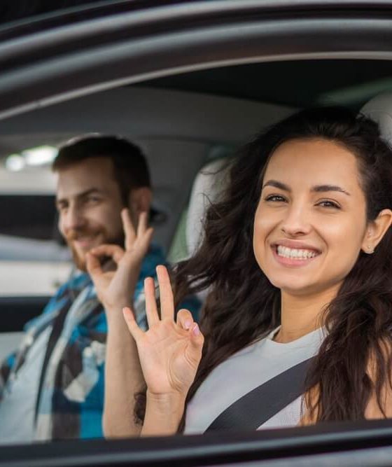 Smiling driver and passenger make “OK” hand signs inside a car, highlighting permissive driving.