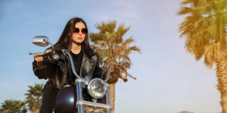 Woman in a black leather jacket and sunglasses posing on a cruiser motorcycle near palm trees at sunset, highlighting motorcycle insurance protecting riders.