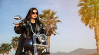 Woman in a black leather jacket and sunglasses posing on a cruiser motorcycle near palm trees at sunset, highlighting motorcycle insurance protecting riders.