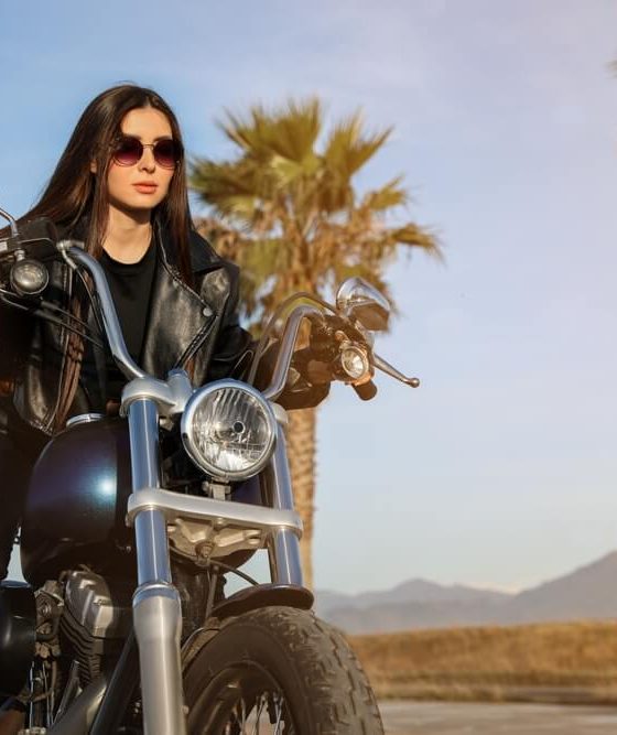 Woman in a black leather jacket and sunglasses posing on a cruiser motorcycle near palm trees at sunset, highlighting motorcycle insurance protecting riders.
