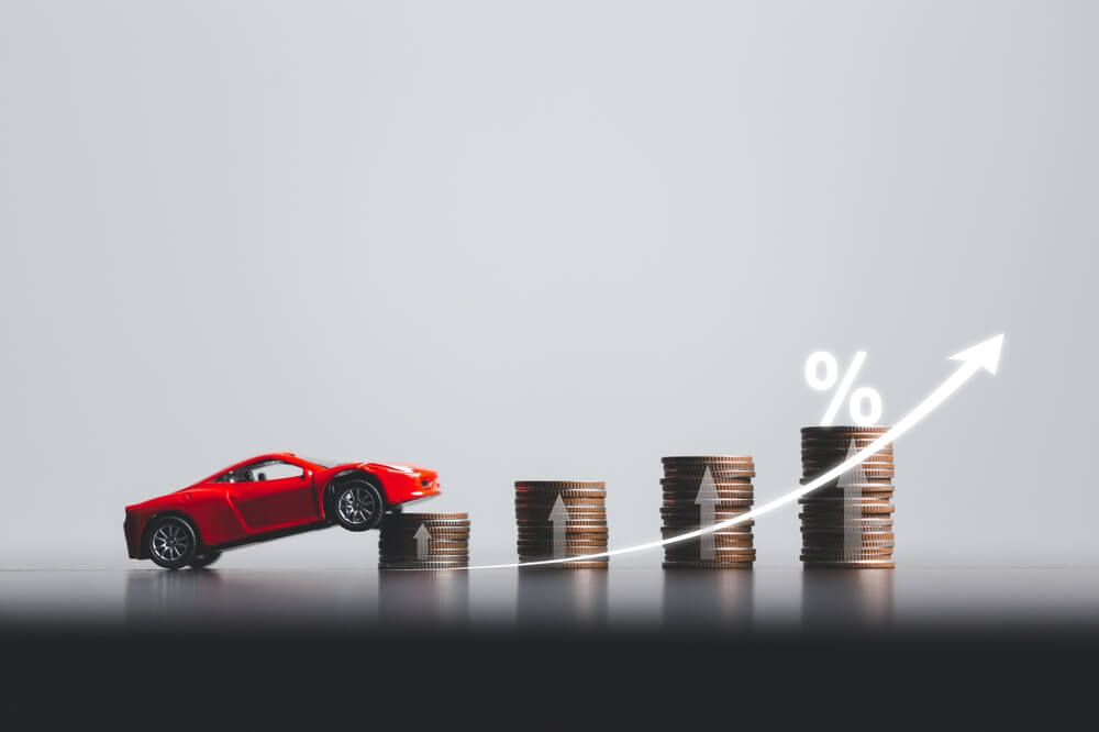 Rising car insurance rates illustrated by a red car climbing stacks of coins with an upward arrow and percentage symbol.