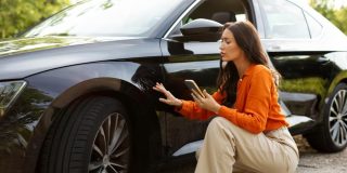 Driver inspecting damage to a black car while using a smartphone after an accident, representing car insurance rates and coverage costs.