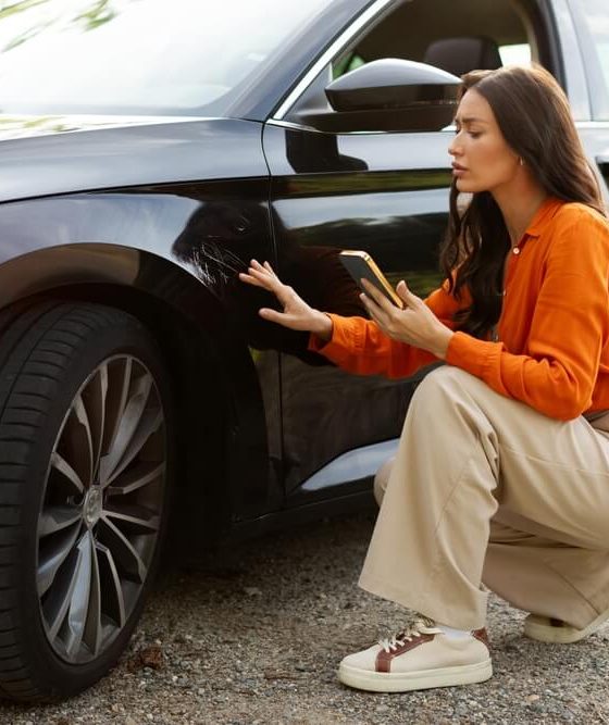 Driver inspecting damage to a black car while using a smartphone after an accident, representing car insurance rates and coverage costs.