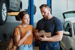 Customer and mechanic review a repair estimate on a clipboard inside an auto shop, showing why car repairs cost more than ever.
