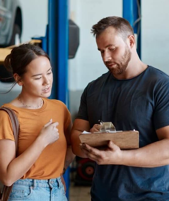 Customer and mechanic review a repair estimate on a clipboard inside an auto shop, showing why car repairs cost more than ever.