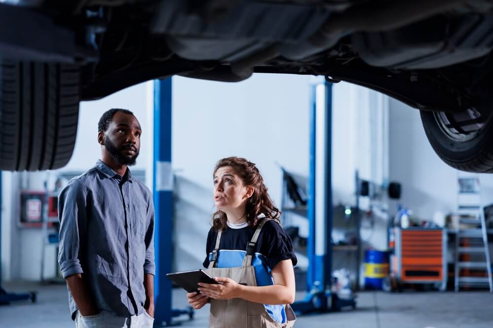 Mechanic explains issues under a lifted car to a customer in an auto repair shop, showing why car repairs cost more than ever.